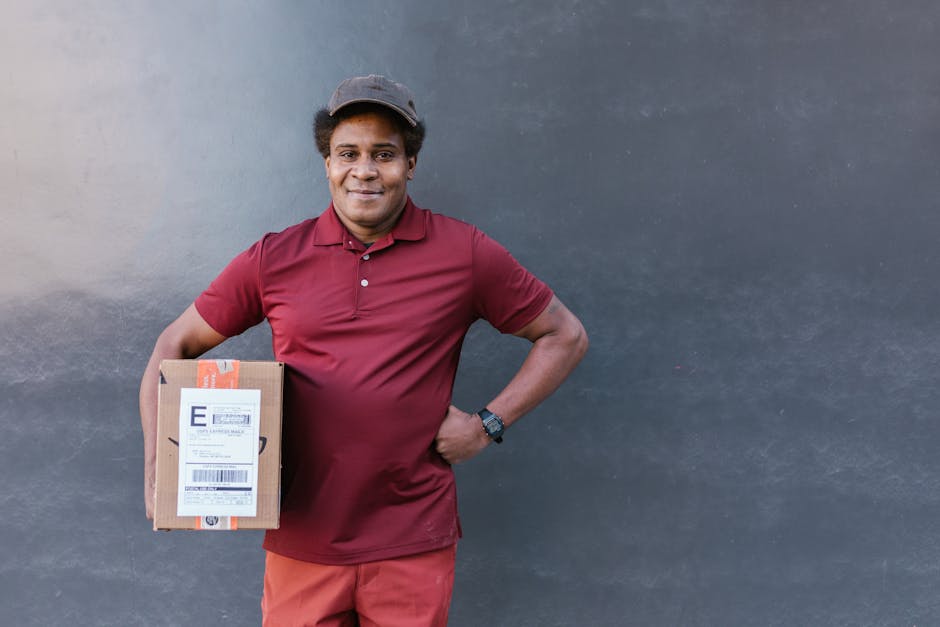 A friendly delivery person with dark skin and curly hair, dressed in a maroon polo shirt and matching cap, is standing against a plain, dark grey wall. He is smiling while holding a small, rectangular