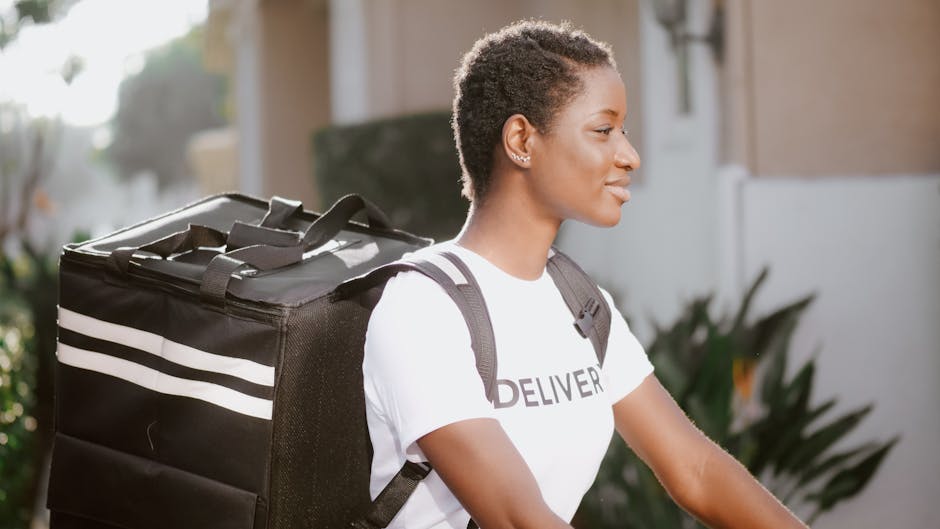 A young woman with short, curly hair and medium skin tone, wearing a white T-shirt with the word 'DELIVERY' partially visible, is carrying a large, black insulated delivery bag with white stripes on h